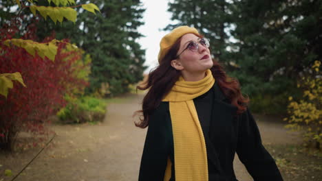 artista con el cabello rojo largo ondulado, con un abrigo negro y boina amarilla, ajusta su cabello mientras camina a través de un parque exuberante, rodeado de colorido follaje de otoño