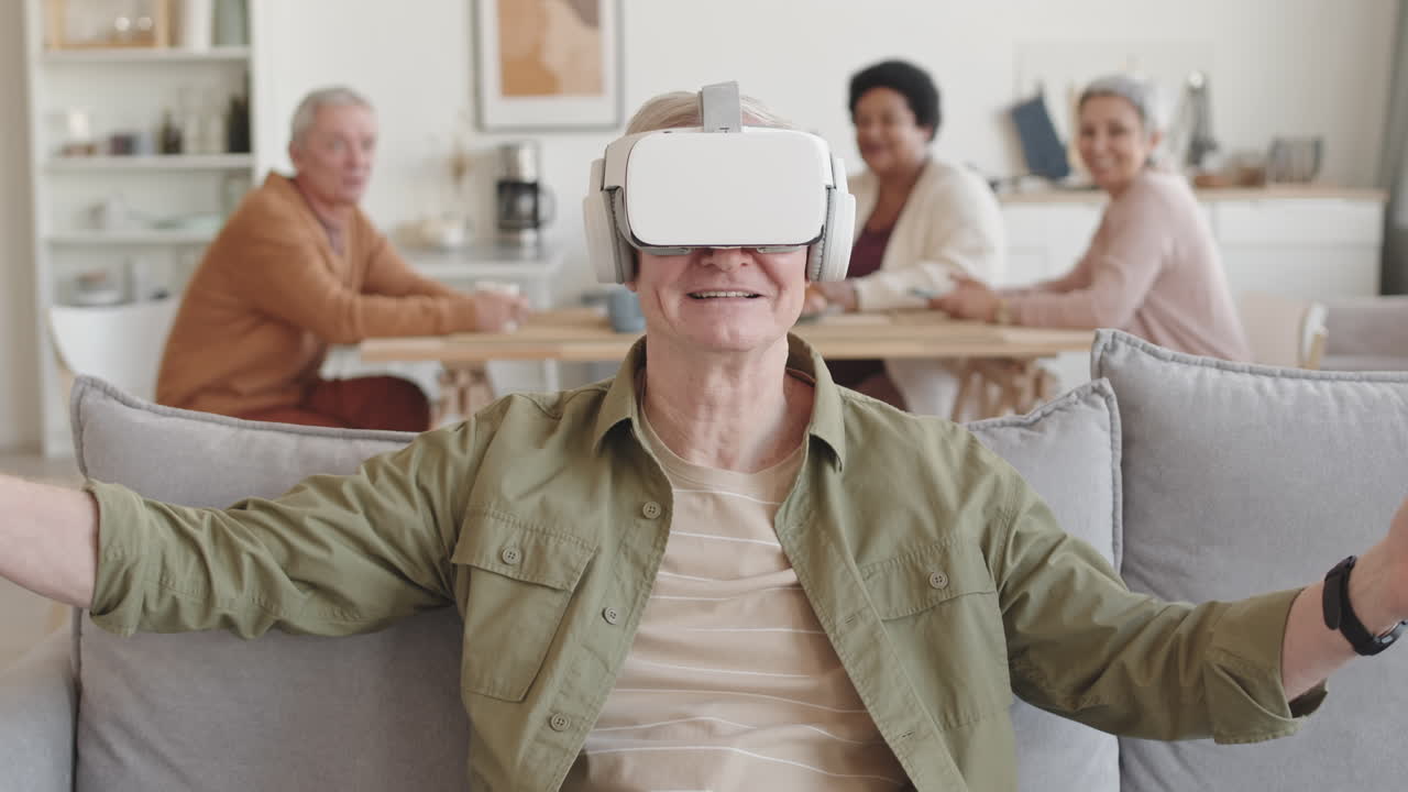 Waist-up of happy senior man wearing virtual reality headset, sitting on couch in living room, raising hands up, talking, smiling