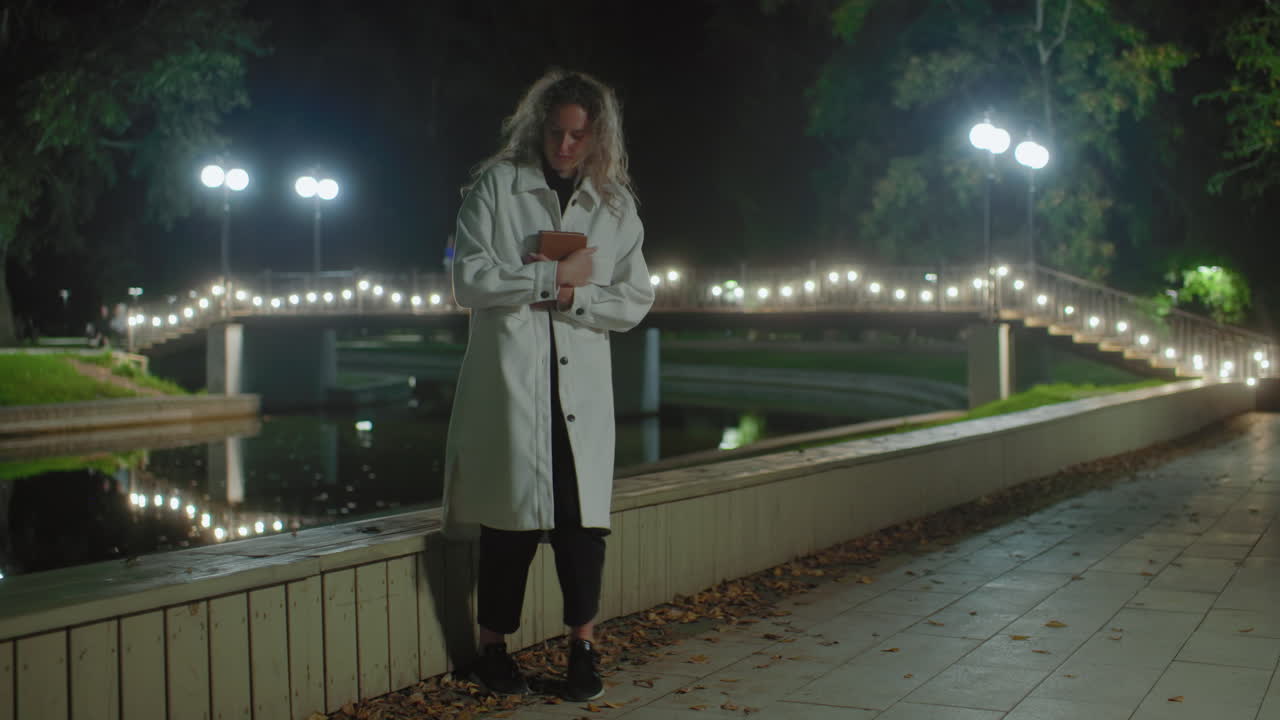 Student in white coat approaches wooden fence near water at night, crosses legs, opens book to read under glowing lamplight, reflection dancing on calm pond