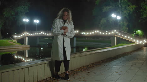student in white coat approaches wooden fence near water at night, crosses legs, opens book to read under glowing lamplight, reflection dancing on calm pond
