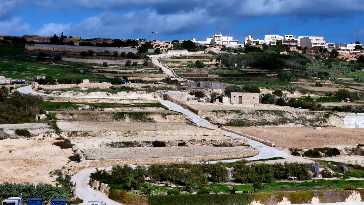 Una vista de gran angular y perspectiva alta captura antiguos campos de terrazas de piedra caliza ascendiendo por una escarpada ladera mediterránea hacia edificios residenciales blancos bajo un cielo azul intenso y nublado durante el día.