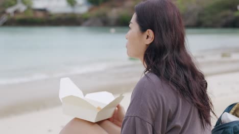 Asian-woman-eating-takeout-food-while-sitting-on-a-beach,-portrait-mid-shot