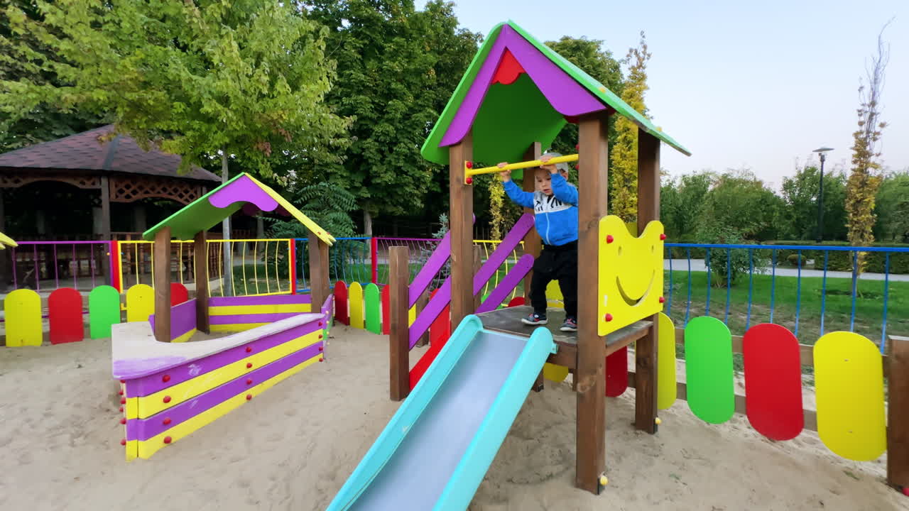 Caucasian toddler having good time at the playground. Cute little boy jumps, hangs on the horizontal bar and goes down by the slide.