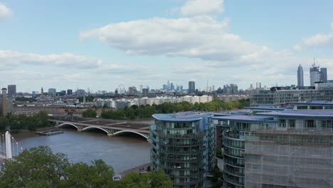 Rising-footage-of-cityscape.-Modern-apartment-houses-at-Thames-riverbank-and-city-panorama-revealing-behind-trees.-Red-train-heading-to-Victoria-station.-London,-UK