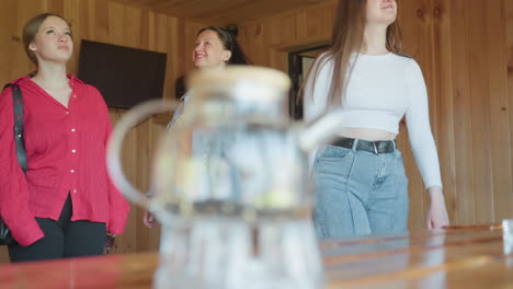 close up of glass jar with tea leaves and candles on wooden table inside cabin as three students walk into bright room in background looking around with interest and excitement