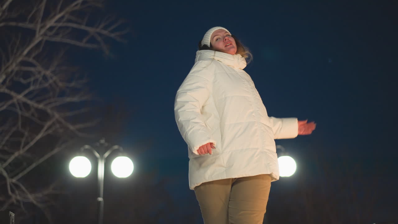 Back view of girl in white puffer coat and headphones swaying gently to music on snowy walkway under glowing lights at night tranquil serene winter park ambiance with graceful motion