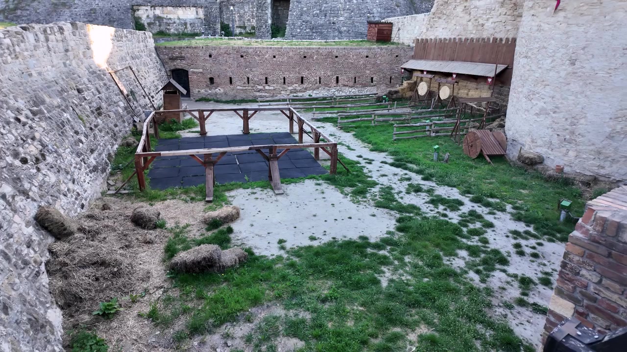 I visitatori possono ammirare le mura in pietra di un'antica fortezza. Nel cortile ci sono aree erbose e una struttura in legno. Il sole splende sulle antiche pietre mentre la natura circonda il sito.