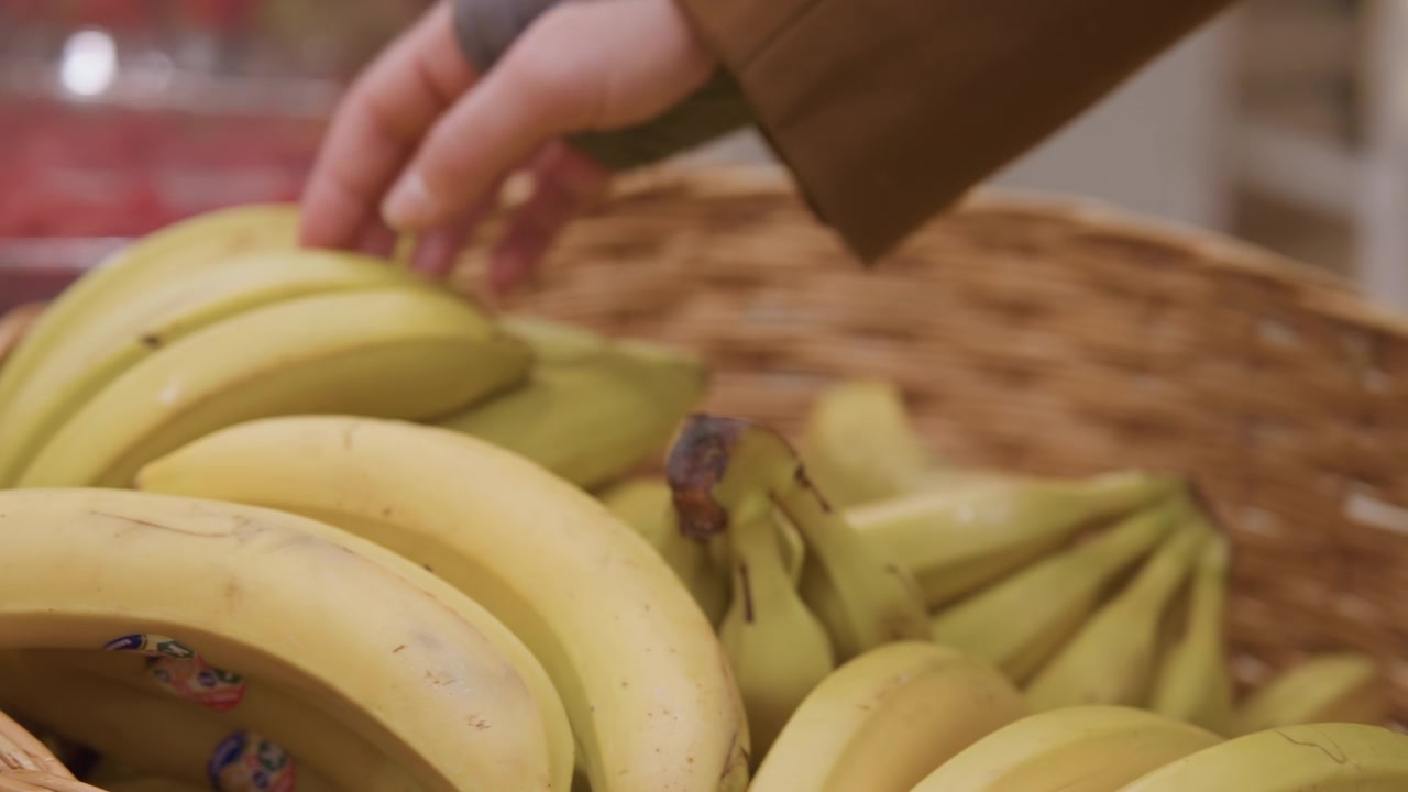 Mano che raccoglie banane mature in un cesto in un mercato coperto, acquirente casuale che indossa un cappotto marrone e guanti senza dita seleziona un grosso casco giallo, illuminazione calda e soffusa, espositore in vimini e pomodori sullo sfondo.