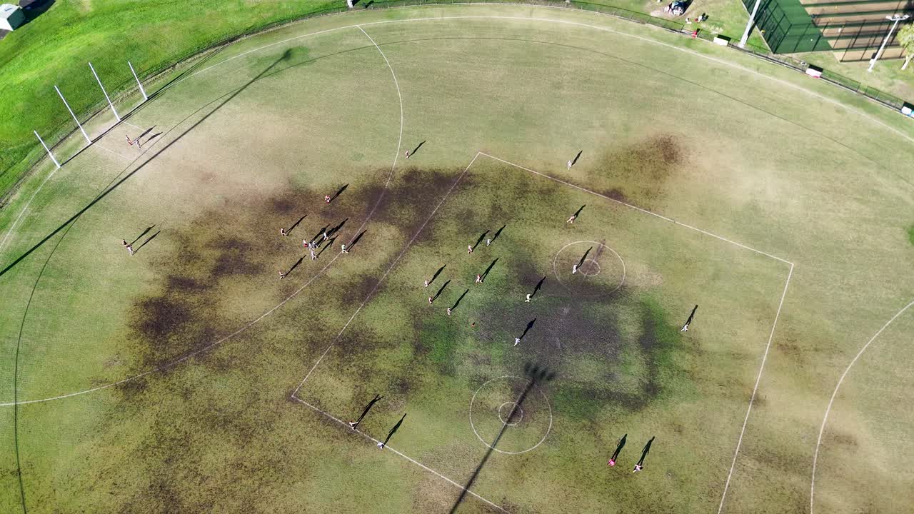 Drone captures an Australian football match on a grassy oval, with long shadows cast by players during sunset