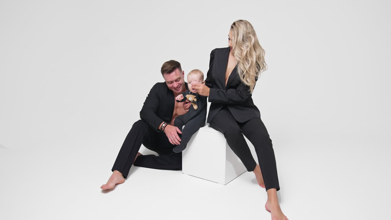 Woman sits on the white cube holding her little son. Smiling father sits beside looking at his baby. White backdrop.
