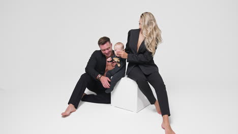 woman sits on the white cube holding her little son. smiling father sits beside looking at his baby. white backdrop.