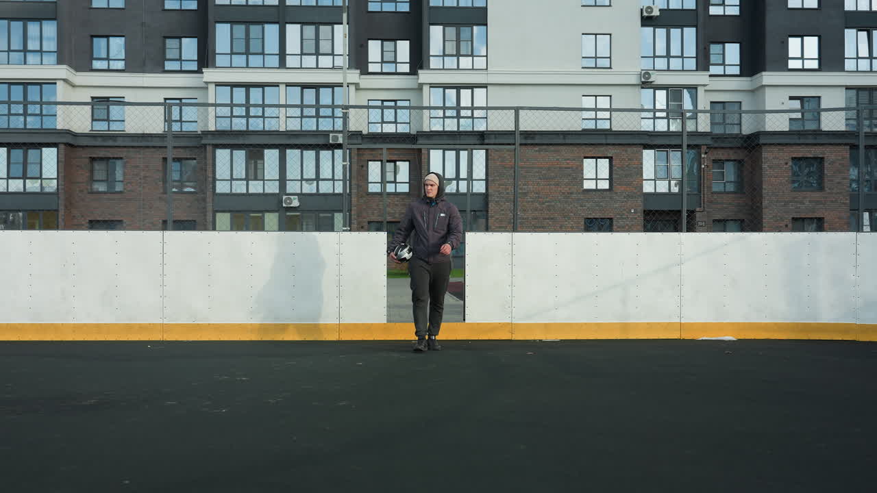 futbolista con capucha y gorra sosteniendo la pelota en la mano derecha camina a lo largo del campo de fútbol con poste de gol en el fondo, edificio residencial alto, y la luz del sol proyectando sombra en el suelo