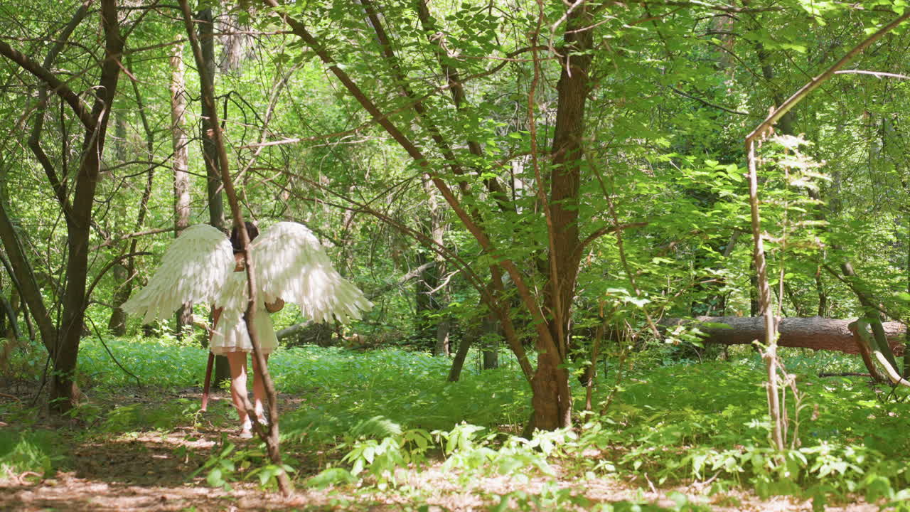 Rear view of graceful lady wearing white wings walking barefoot into bright green forest, surrounded by sunlight filtering through trees, symbolizing freedom and peace