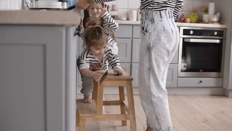 young stylish mother helps daughter turn the pancake with a shovel and having fun while cooking together in kitchen at home with two children