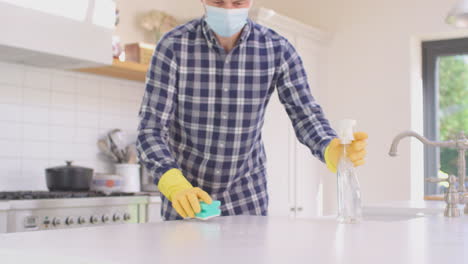 man in face mask at home in kitchen wearing rubber gloves cleaning down work surface using cleaning spray during health pandemic  - shot in slow motion