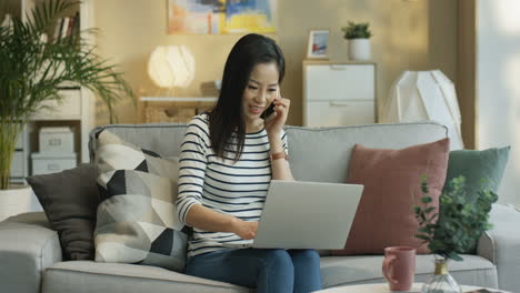 young woman wearing striped blouse with a laptop on laps and talking cheerfully on the phone