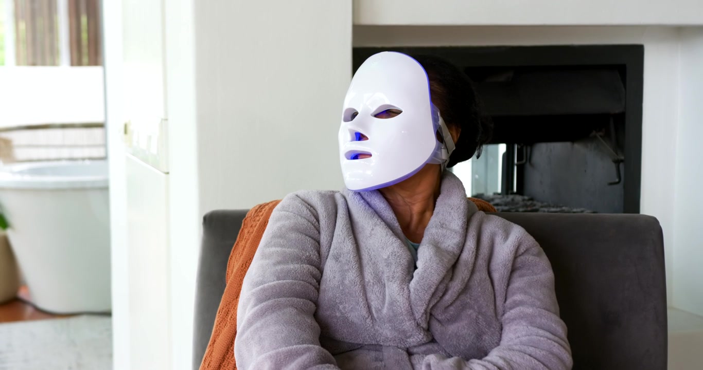 Mulher afro-americana idosa de roupão virando a cabeça em casa enquanto máscara de LED é ativada para terapia