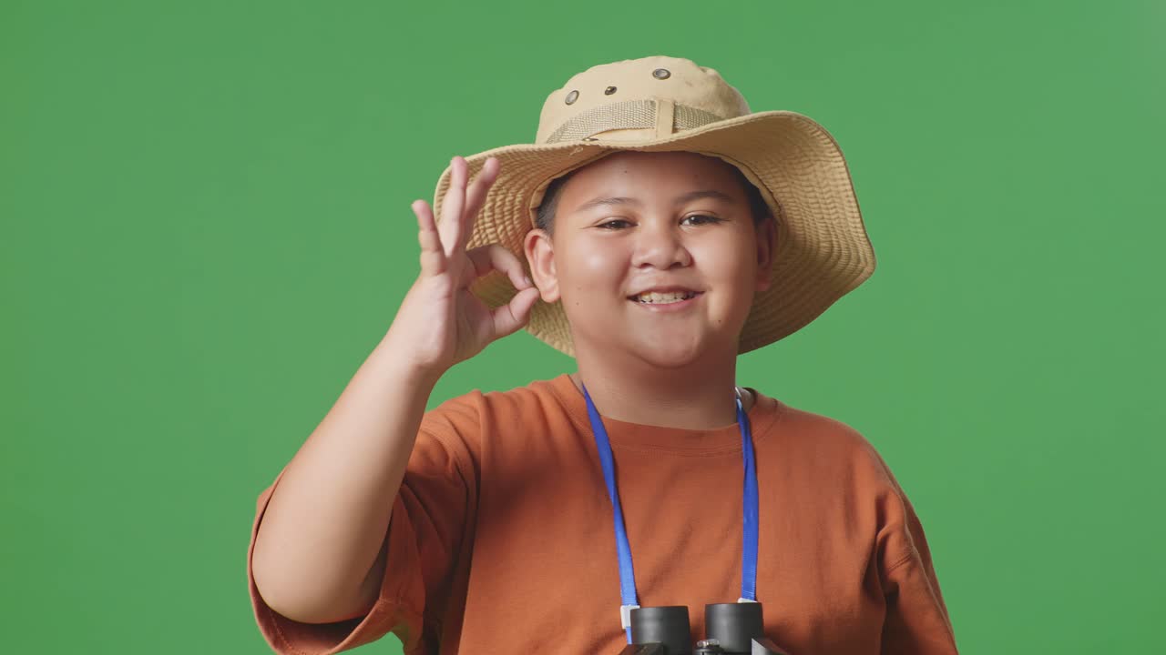 모자를 입은 아시아 관광 소년은 안경을 통해 쳐다본 후 은 몸짓을 보여줍니다. 소년 연구자는 녹색 화면 배경에서 무언가를 조사합니다. 여행 관광 모험 개념, 근접