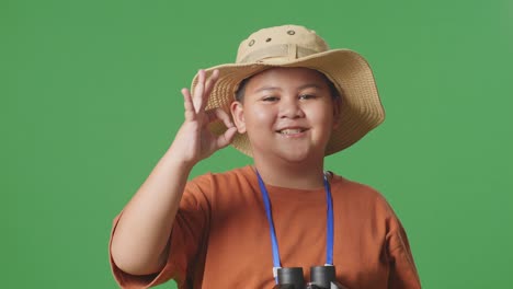 asian tourist boy with a hat showing okay gesture after looking through the binoculars. boy researcher examines something on the green screen background, travel tourism adventure concept, close up