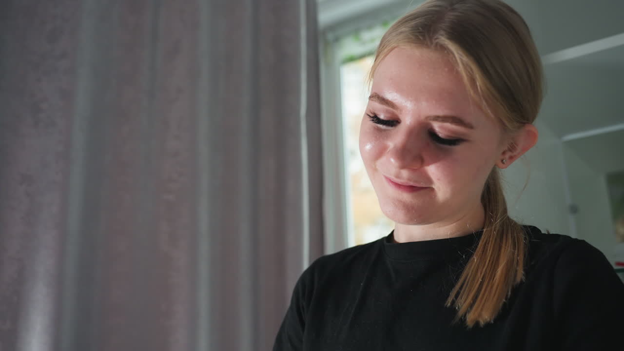 Close up of young female massage therapist smiling softly while concentrating on task, with blurred background showing cabinet and soft curtains, natural daylight illuminating peaceful expression