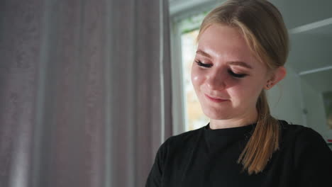 close up of young female massage therapist smiling softly while concentrating on task, with blurred background showing cabinet and soft curtains, natural daylight illuminating peaceful expression