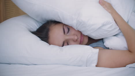 young woman covering ears with a pillow. annoyed by loud neighbors can not sleep. irritated white girl with insomnia wakes up.