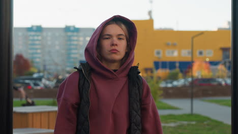 portrait wellness minded woman in burgundy hoodie and black vest posing with serious facial expression in outdoor gym park featuring blurred pedestrians foliage conveying focus confidence strength