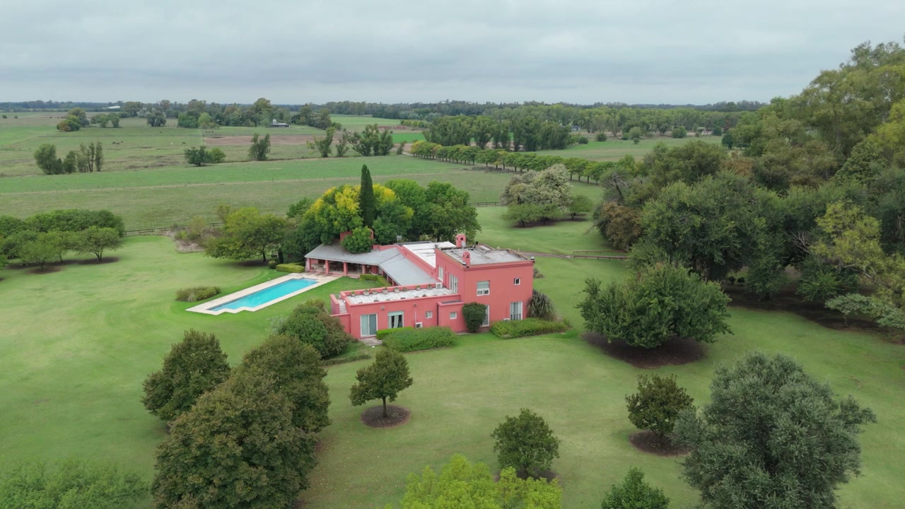 Vista aerea si avvicina a una casa di campagna rosa con piscina, circondata da campi verdi e alberi.