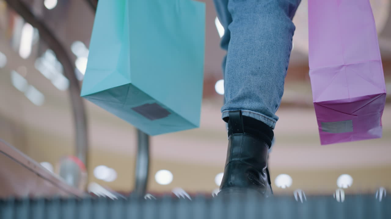진즈와 부츠를 입은 사람이 움직이는 에스컬레이터에서 분홍색과 민트 쇼핑 가방을 들고 배경에 보케 조명과 함께 나가는 아래쪽 몸의 근접한 모습