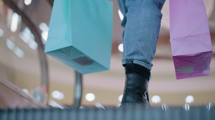 Primo piano della parte inferiore del corpo di una persona in jeans e stivali che trasporta borse della spesa rosa e verde menta su una scala mobile, mentre esce con luci bokeh sullo sfondo