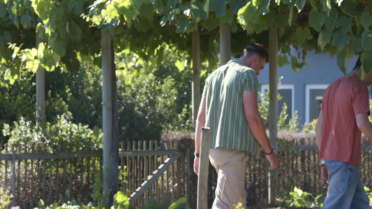 パーゴラの下の庭の小道を歩く十代の少年と成人の男性。成人が誘導するように手を置き、十代の少年が先導している。