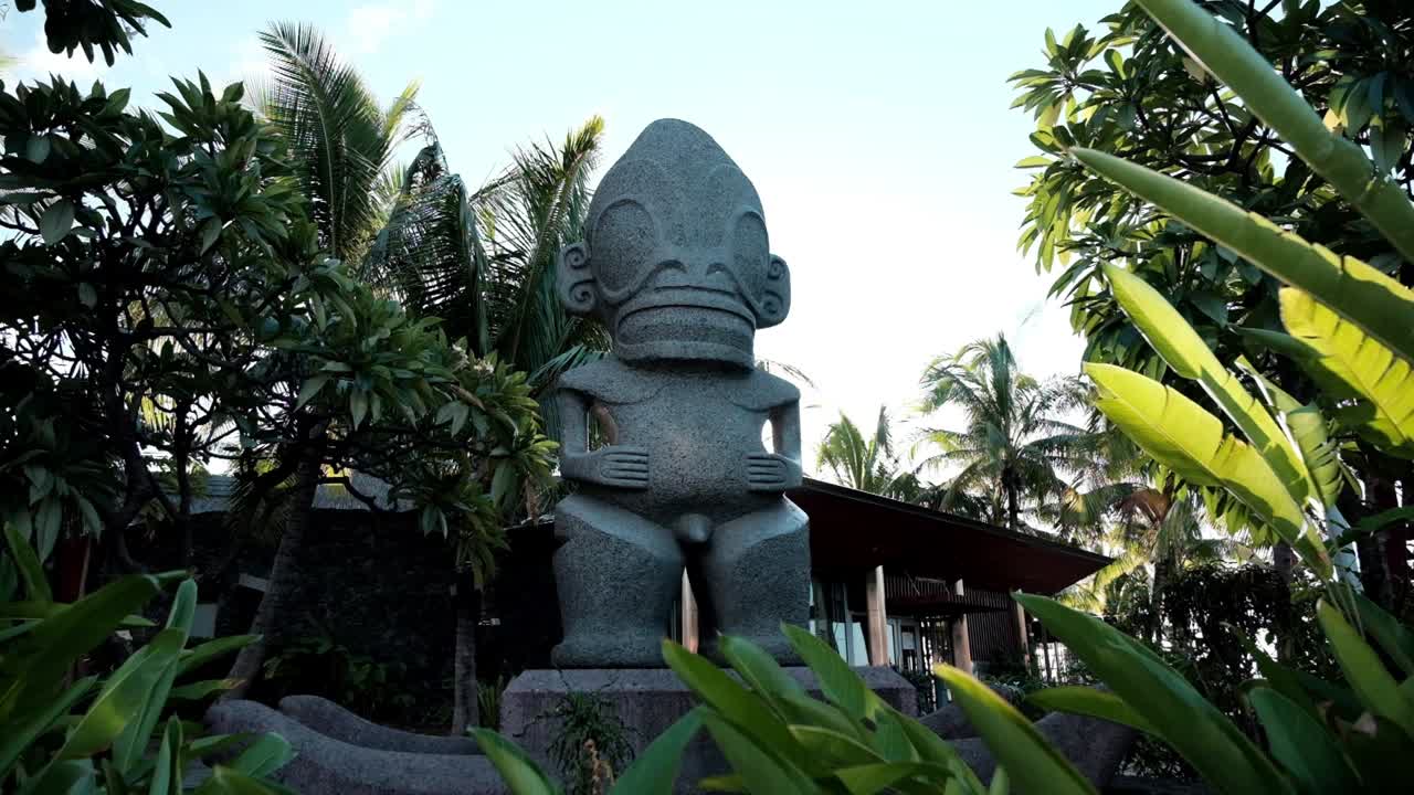gran estatua de tiki de hormigón tallado en papetee tahiti rodeada de plantas tropicales y palmeras con una llamarada de luz brillante al final