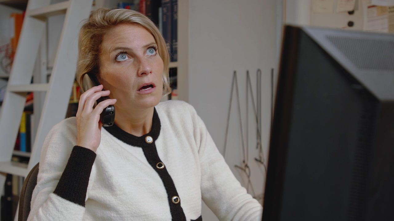 Ung profesjonell forretningskvinne jobber på datamaskinen sin ved skrivebordet på et hjemmekontor mens hun besvarer en irriterende telefonsamtale.