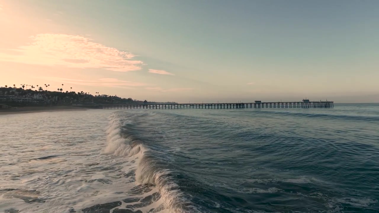 Volo mozzafiato di un drone cinematografico che plana appena sopra le onde all'alba, viaggiando lungo la costa della California verso l'iconico molo di San Clemente.