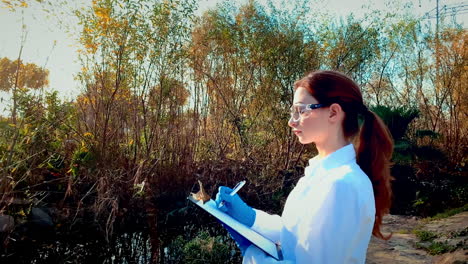 A-young-woman-scientist-at-a-creek,-wearing-protective-eyewear-and-a-lab-coat,-taking-notes-on-a-clipboard