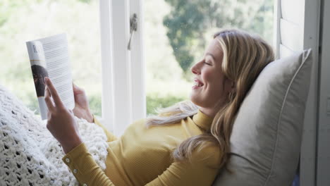 happy biracial woman sitting next to window reading book at home, slow motion