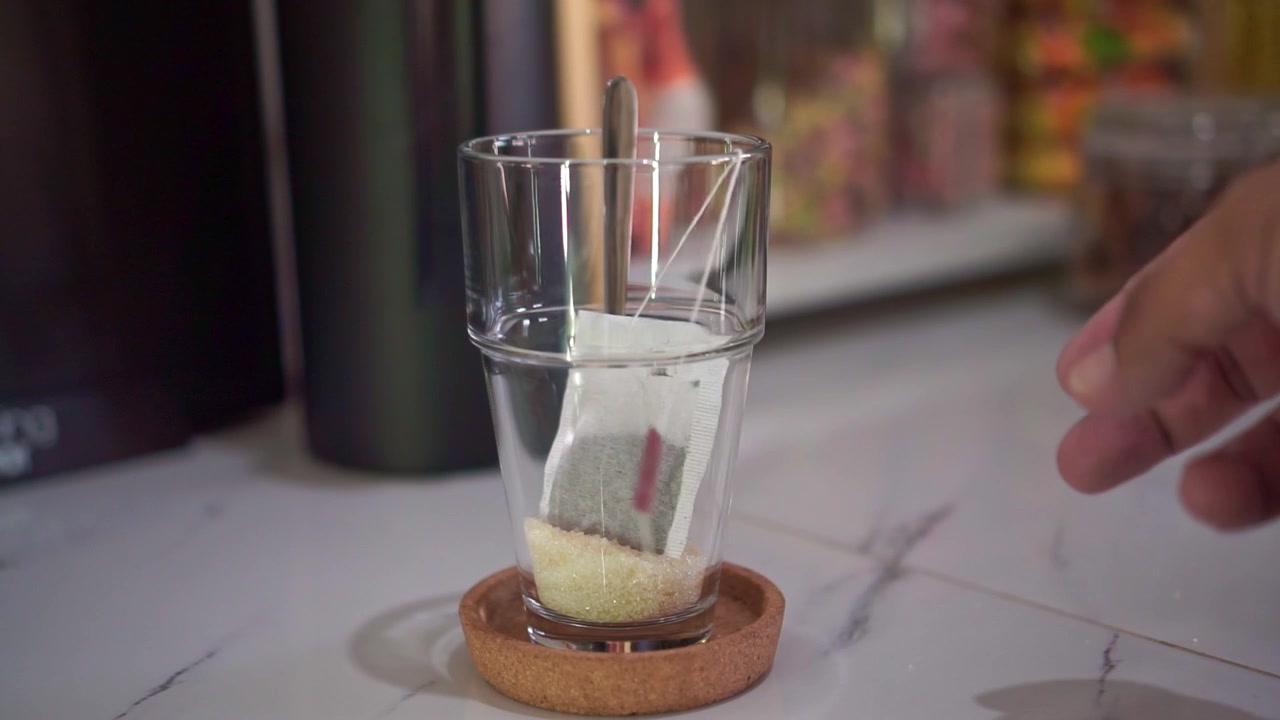 Bustina di tè che cade in una tazza di vetro con cucchiaino e zucchero all'interno. Preparazione casalinga del tè, concetto di bevanda calda, filmato stock culinario