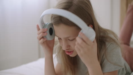 una niña está poniendo auriculares inalámbricos y tomando un teléfono inteligente para escuchar música y usar internet acostada en la cama