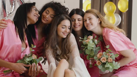 group of multiethnic female friends caressing bride who is wearing white silk nightdresses sitting on bed holding bouquets decorated with word 'bride' ballons in bridal gathering