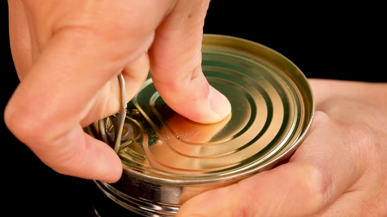 A hand opens a metal can of spaghetti using a pull tab, revealing pasta in tomato sauce. Close-up, well-lit, black background, steady camera