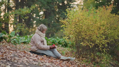 woman wearing scarf and puffer jacket sits cross-legged on dry autumn leaves near colorful bush, holding small book in peaceful forest setting, absorbed in reading during quiet moment in nature