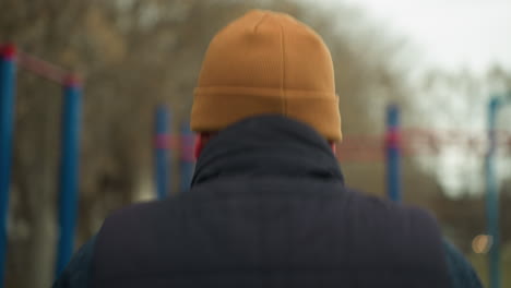 vista trasera de un entrenador con una gorra marrón corriendo rápido en un gimnasio al aire libre, con una vista borrosa de un coche pasando y barras de hierro rojas y azules en el fondo