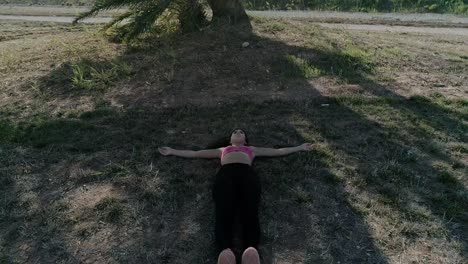 drone shot of young brunette woman lying on the ground next to a big tree