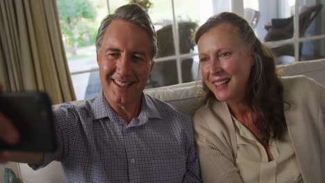 feliz pareja caucásica mayor sentada en la sala de estar sonriendo y tomando una selfie
