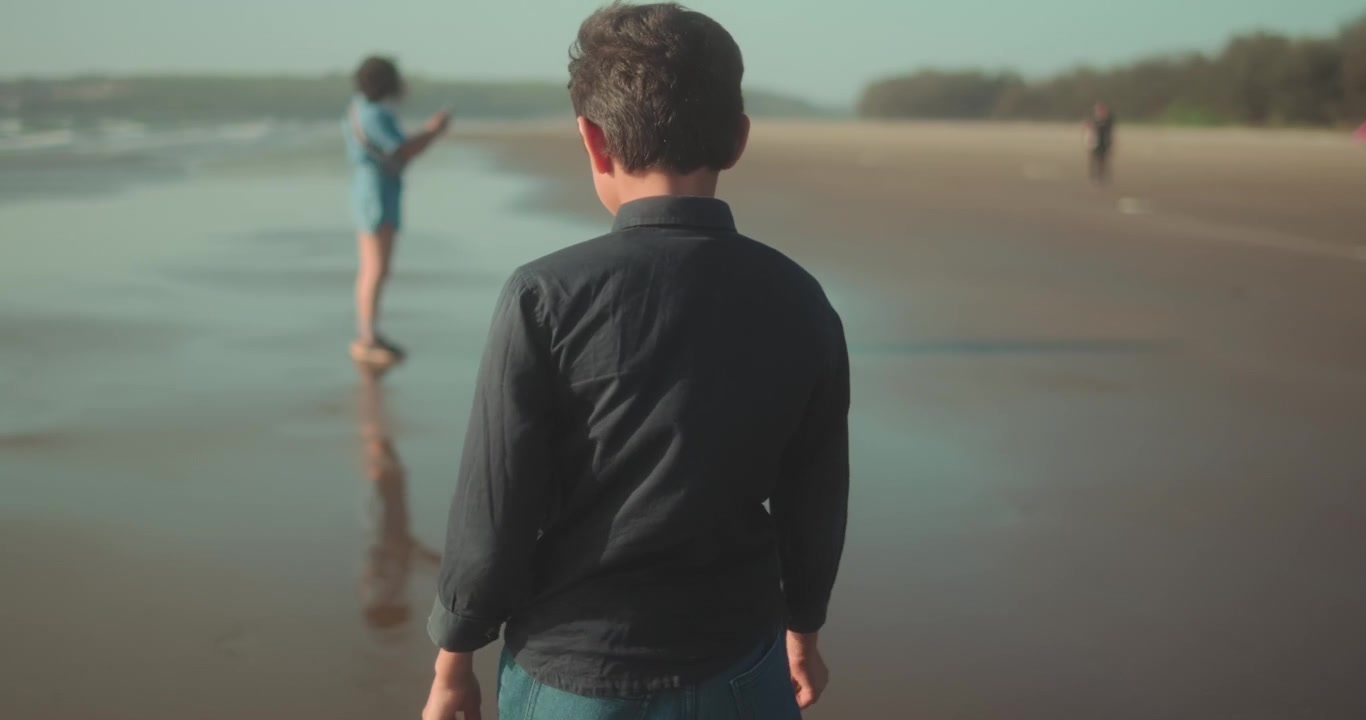 Giovane ragazzo che cammina lungo la spiaggia sabbiosa in riva all'oceano durante una calda vacanza estiva, esplorando l'acqua e riflettendo su una figura distante