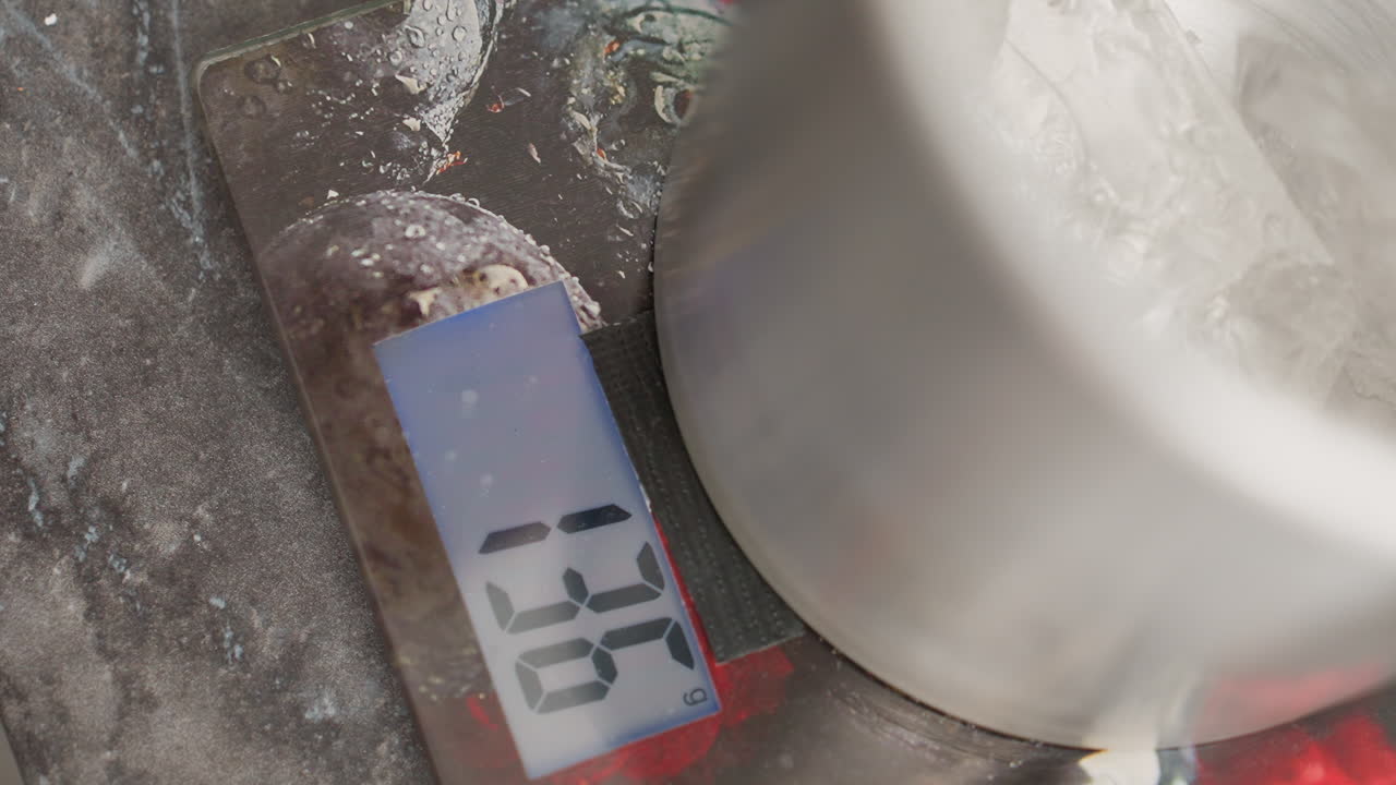 Close up of digital kitchen scale displaying weight reading 142 grams as translucent rubber-like substance is being dropped into metal container placed on scale
