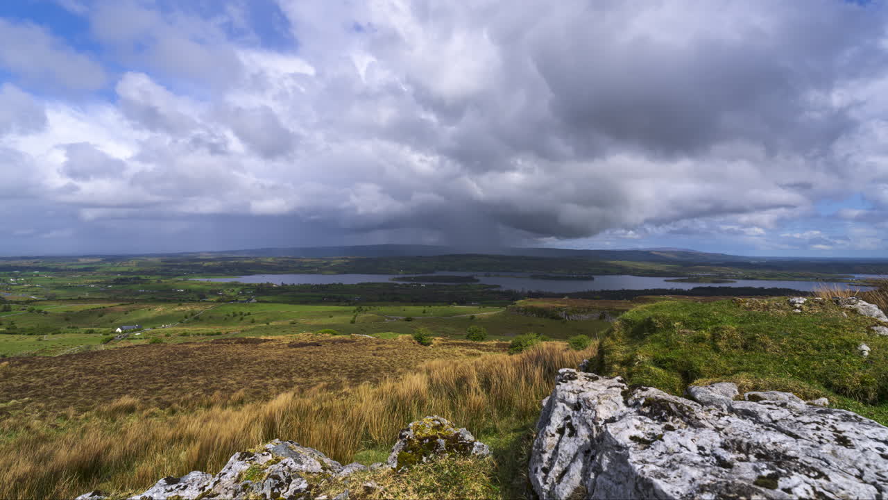 Timelapse af et landligt landskab med en dal og en stenblok i forgrunden samt et passerende regnvejr, bakker og en sø i det fjerne på en forårsdag set fra Carrowkeel i County Sligo i Irland