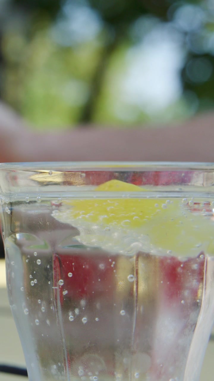 Hand drops cherry into glass of sparkling water with lemon, daylight, shallow depth of field