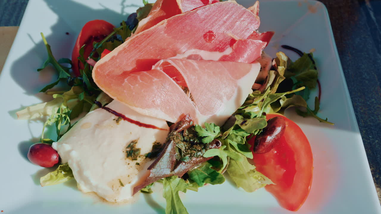 Close up of a gourmet salad with fresh mozzarella, cured meat slices, tomatoes, olives and mixed greens on a white plate in bright daylight