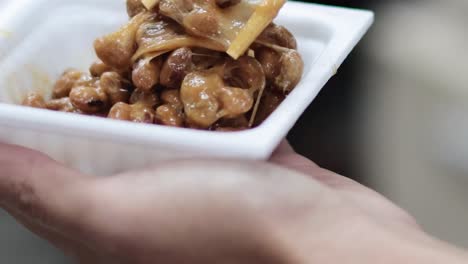 Close-up of sticky natto being lifted with chopsticks from a small tray, showcasing its stringy texture.